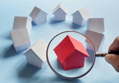 Betrachtung einer Immobilie: Ein rotes Haus wird mit der Lupe angesehen unter mehreren Häusern in einem Kreis.